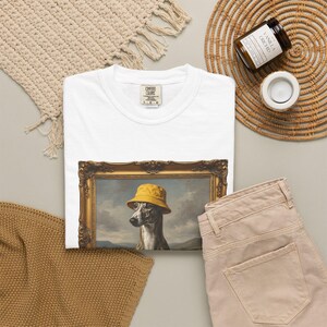 Może przedstawiać: Biały t-shirt z nadrukiem graficznym przedstawiającym charta w żółtym kapeluszu typu bucket hat, umieszczonym w ozdobnej złotej prostokątnej ramie. Koszulka posiada metkę marki Comfort Colors przy wewnętrznym dekolcie z nadrukowanym rozmiarem L-G-G.