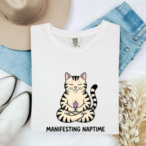 May include: A white t-shirt featuring a graphic of a cream-colored tabby cat with dark stripes sitting in a meditative pose while holding a purple crystal. Below the cat, the text reads "MANIFESTING NAPTIME" in bold, black, capital letters.