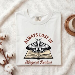 May include: A white short-sleeve t-shirt featuring a graphic of an open book with a moth resting on top. The text "ALWAYS LOST IN" is printed in a rust-colored arch above the moth, and "Magical Realms" is printed below in the same color.