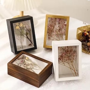 May include: Four rectangular shadow boxes with dried flowers inside. The frames are black, gold, white, and brown. The boxes are displayed on a white surface. The flowers are various shades of beige, pink, and brown.