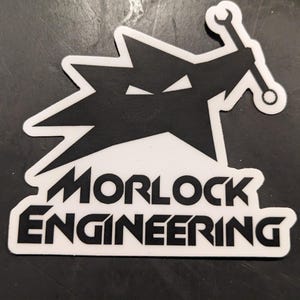 May include: A white sticker with a black graphic of a stylized wolf head holding a wrench. The text "MORLOCK ENGINEERING" is printed in bold, black letters below the graphic. The sticker has a white border.