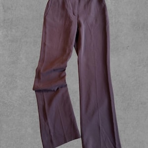 Può includere: Un paio di pantaloni marrone scuro a gamba larga. I pantaloni hanno un design classico con chiusura a bottone e cerniera, passanti per cintura e tasche anteriori. Il tessuto sembra essere un materiale liscio e tessuto.