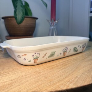 Könnte beinhalten: Eine rechteckige, cremefarbene Auflaufform mit einem dekorativen Gartenmotiv. Der Rand zeigt Illustrationen von Topfblumen, Gartengeräten und Libellen. Die Form steht auf einem Holztisch.