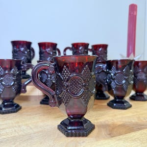 Op de afbeelding: Een witte keramische mok met een zwarte handgreep en de tekst "But First, Coffee" in een vetgedrukte, zwarte letter. De mok staat tegen een vage achtergrond, wat een gezellige omgeving suggereert.