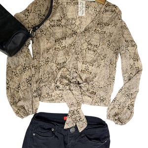 Peut inclure: Un chemisier à manches longues avec un imprimé peau de serpent beige et marron, doté d'un col en V, d'une fermeture boutonnée sur le devant et d'un ourlet à nouer. Il est accompagné d'un pantalon noir et d'un sac à bandoulière noir.