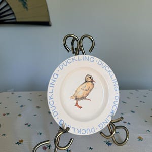 May include: A small, white plate with a light blue border and the word "DUCKLING" printed around the edge. The plate features a watercolor illustration of a yellow duckling with orange feet. The plate is displayed on a gold-colored metal stand.