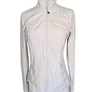 Op de afbeelding: Een lichtbeige sportjack met een hoge kraag en een ritssluiting over de hele lengte. De jas heeft lange mouwen, twee zakken met ritssluiting en een getailleerde pasvorm. De jas wordt op een mannequin getoond.