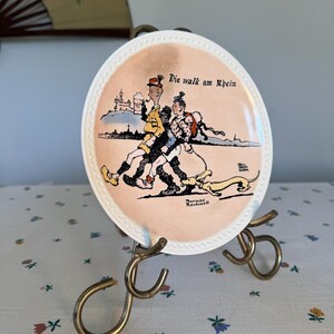 Pode incluir: Um prato decorativo vintage com um centro rosa apresentando uma ilustração de desenho animado de pessoas andando com um cachorro. O prato tem uma borda branca e é exibido em um suporte de latão. O texto no prato diz "Die walk am Rhein".
