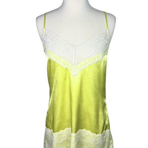 May include: A bright lime green camisole featuring a V-neckline with white floral lace trim. The garment has thin spaghetti straps and a matching white floral lace border along the bottom hem, crafted from a smooth, satin-like fabric.