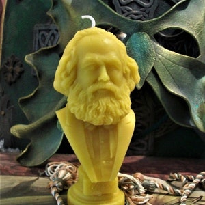 May include: A yellow wax candle shaped like a bust of a man with a beard. The candle is on a small round base and has a wick sticking out of the top.