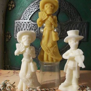 May include: Three decorative figurines made of wax, each depicting a person in a different pose. The figurine on the left is wearing a white dress and holding a fan. The figurine in the center is wearing a yellow dress and holding a fan. The figurine on the right is wearing a black suit and holding a musical instrument.