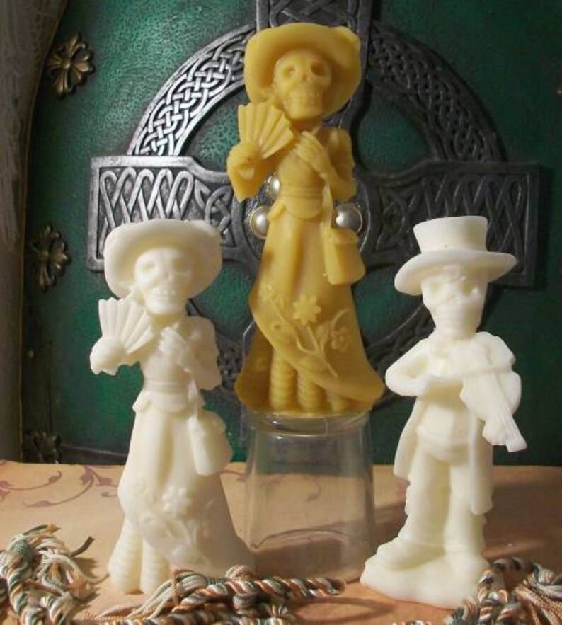 May include: Three decorative figurines made of wax, each depicting a person in a different pose. The figurine on the left is wearing a white dress and holding a fan. The figurine in the center is wearing a yellow dress and holding a fan. The figurine on the right is wearing a black suit and holding a musical instrument.