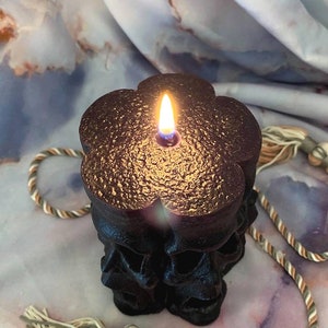 May include: Black skull-shaped candle with a gold metallic finish, lit and burning with a flame. The candle is on a white and brown marble surface with a beige and brown rope with tassels.