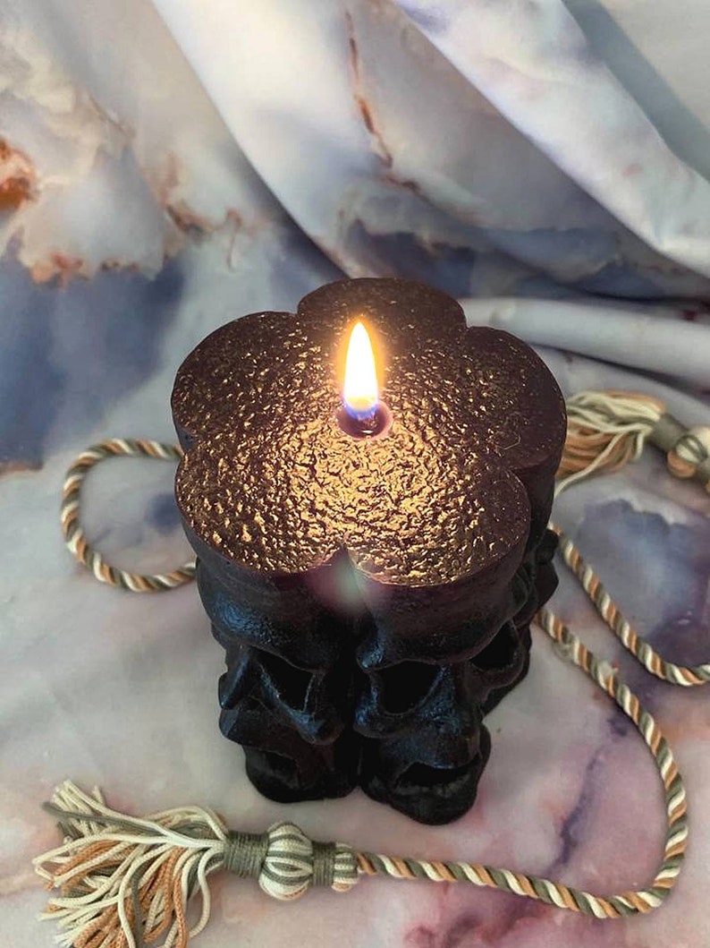 May include: Black skull-shaped candle with a gold metallic finish, lit and burning with a flame. The candle is on a white and brown marble surface with a beige and brown rope with tassels.