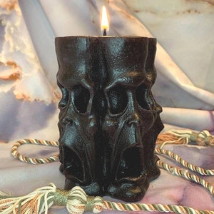 May include: A black candle with a single flame, shaped like three screaming faces. The candle is sitting on a white and brown fabric surface.