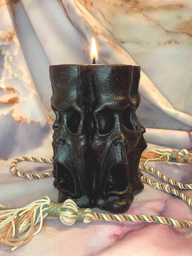 May include: A black candle with a single flame, shaped like three screaming faces. The candle is sitting on a white and brown fabric surface.