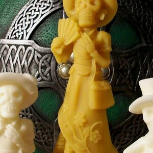 May include: A yellow wax figurine of a skeleton wearing a hat and dress. The figurine is holding a fan and a purse. The figurine is standing on a clear glass base.