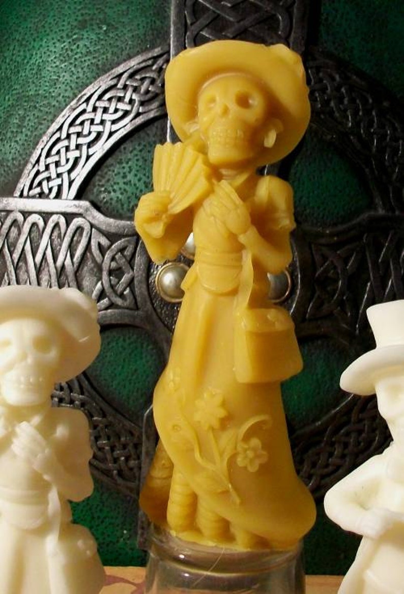 May include: A yellow wax figurine of a skeleton wearing a hat and dress. The figurine is holding a fan and a purse. The figurine is standing on a clear glass base.