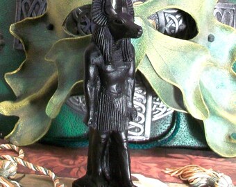 May include: A black statue of the Egyptian deity Anubis, depicted with a jackal head and human body, standing on a rectangular base. The figure wears a traditional pleated kilt and has pointed ears, with a smooth, dark finish throughout the form.