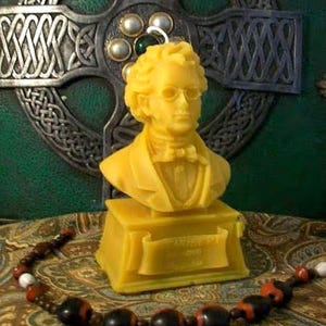 May include: A yellow beeswax candle shaped like a bust of a man with a mustache and glasses. The candle is on a yellow base with the text "W.B. YEATS 1865-1939" on a scroll.