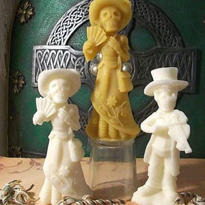 May include: Three white and yellow wax figurines of skeletons dressed in traditional Mexican clothing. The figurines are standing on a table with a green background.