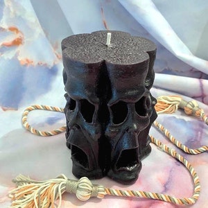 May include: A black pillar candle with a design of multiple faces. The candle has a white wick and is sitting on a white and orange patterned surface.