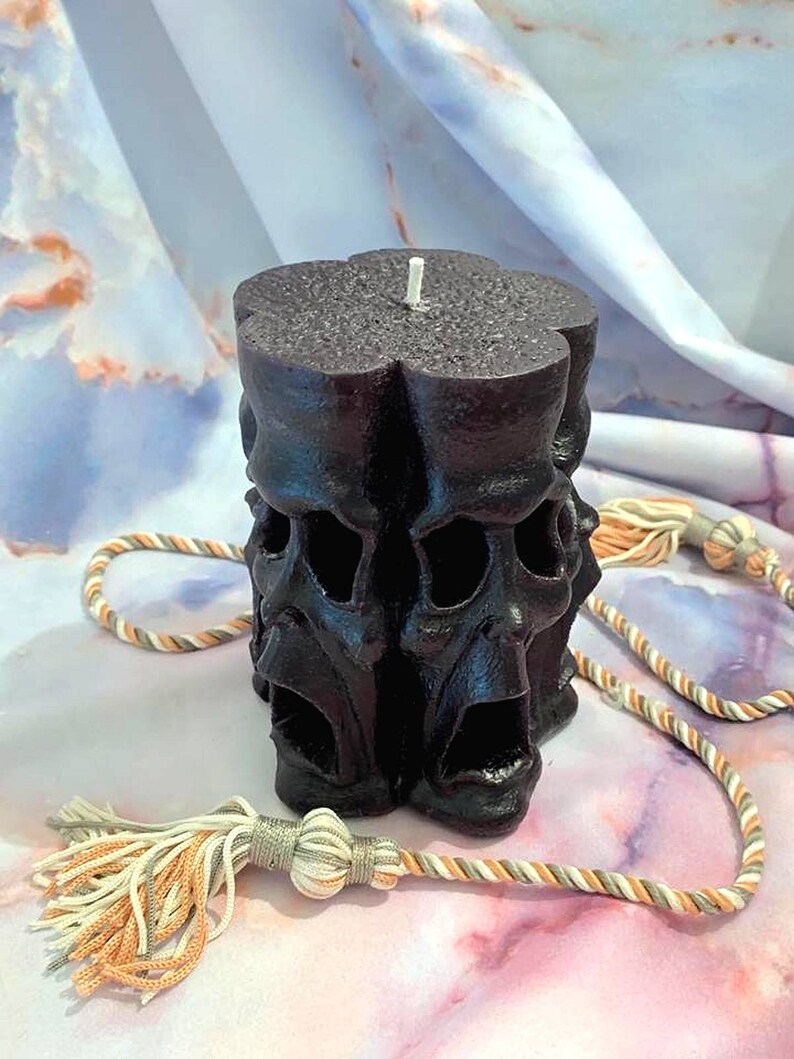 May include: A black pillar candle with a design of multiple faces. The candle has a white wick and is sitting on a white and orange patterned surface.
