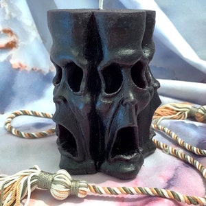 May include: A black pillar candle with three screaming faces carved into the side. The candle is sitting on a white and brown fabric surface.