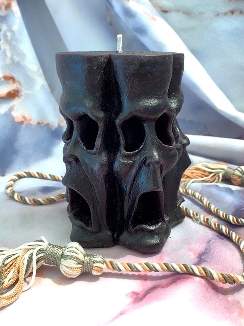 May include: A black pillar candle with three screaming faces carved into the side. The candle is sitting on a white and brown fabric surface.