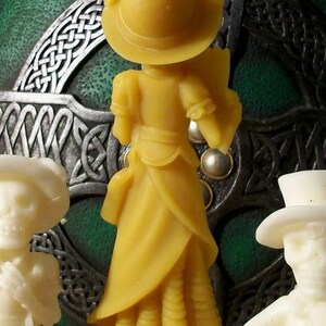 May include: A yellow wax figurine of a woman wearing a hat and a long dress. The figurine is standing on a clear glass base. The background is a green and silver Celtic knot design.