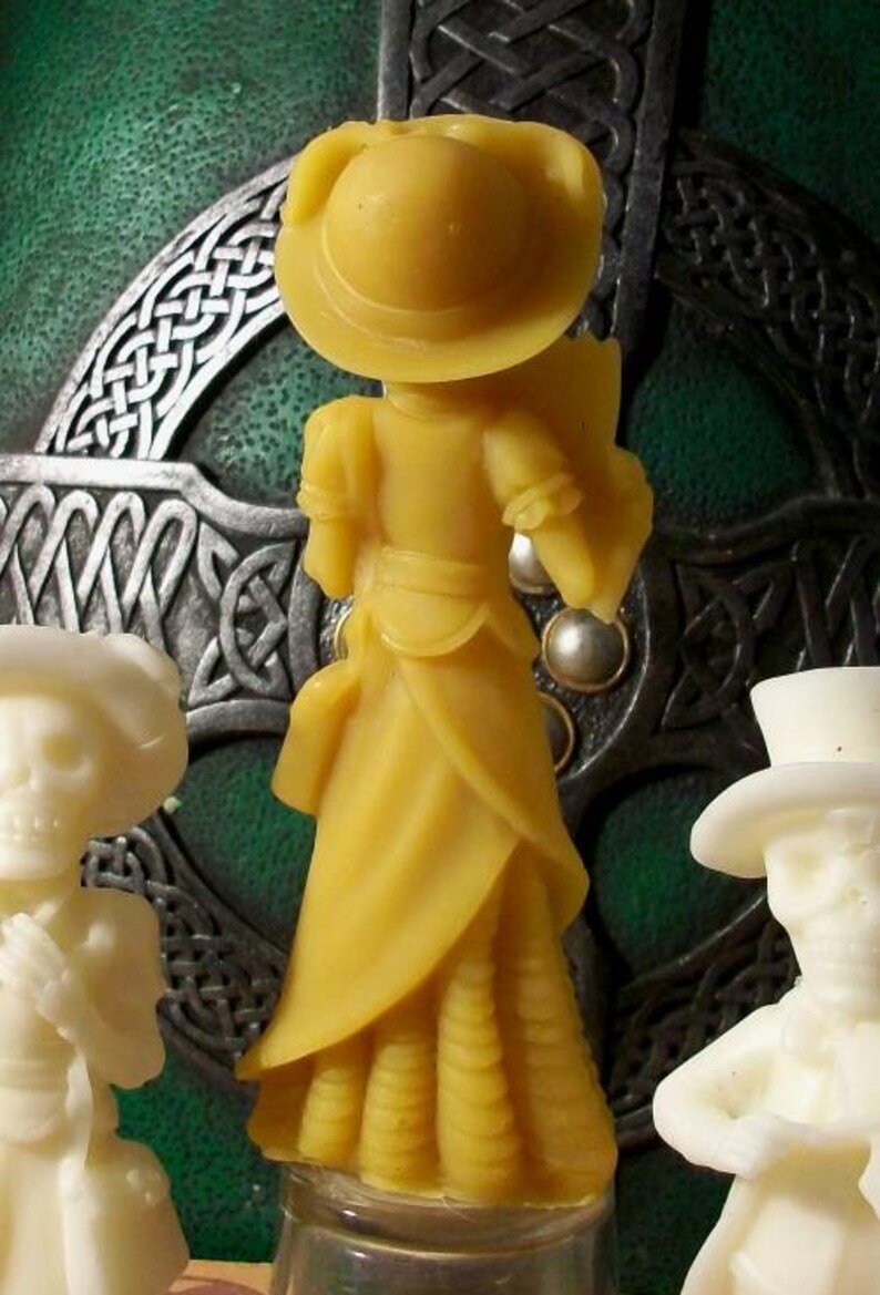May include: A yellow wax figurine of a woman wearing a hat and a long dress. The figurine is standing on a clear glass base. The background is a green and silver Celtic knot design.