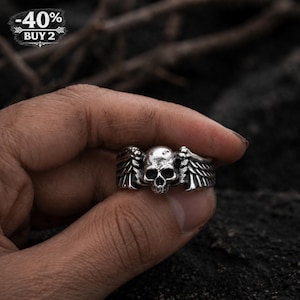 May include: A silver-colored ring featuring a skull with feathered wings. The ring is held in a hand against a dark background. The top left corner of the image has a white and black text that says "-40% BUY 2".