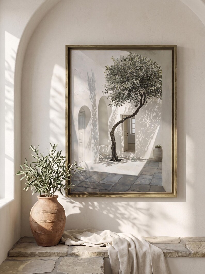 May include: A framed painting depicts a sunlit courtyard with a tree. The artwork features a gold frame, white walls, and a stone floor. A terracotta vase with greenery sits in the foreground. The scene evokes a Mediterranean aesthetic.