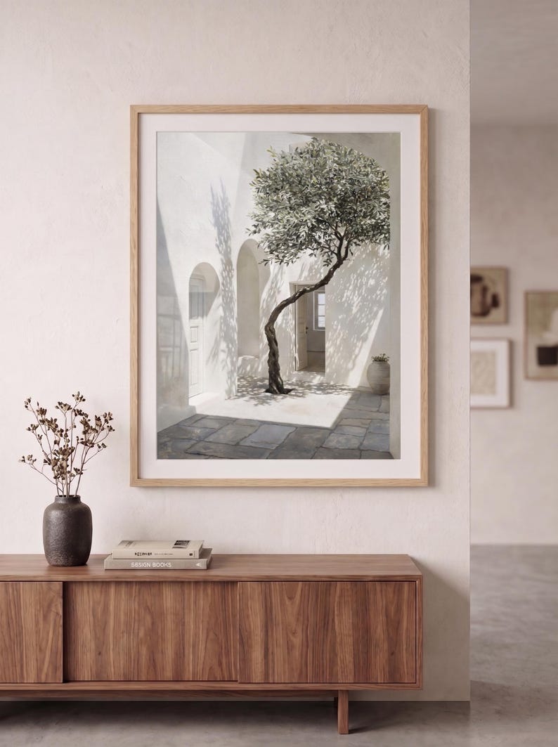 May include: A framed art print depicts a sunlit courtyard with a tree. The artwork features a white building with arched doorways and a stone-paved floor. The tree's branches cast shadows. A dark vase with dried flowers sits on a wooden cabinet.