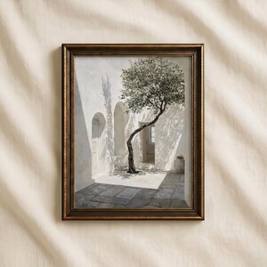 May include: Framed art print depicting a sunlit courtyard with a slender tree. The artwork features a white building with arched doorways and a stone-paved floor. The frame is a dark, warm brown.