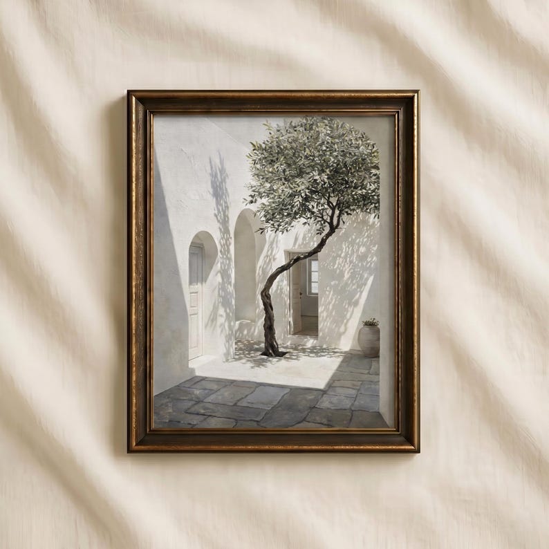 May include: Framed art print depicting a sunlit courtyard with a slender tree. The artwork features a white building with arched doorways and a stone-paved floor. The frame is a dark, warm brown.