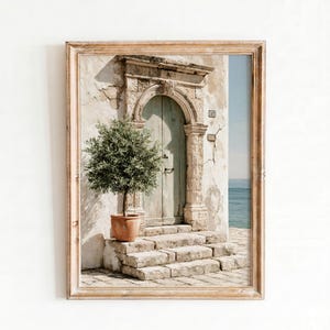 May include: A framed wall art print depicting a weathered stone building with an arched doorway, a potted olive tree on stone steps, and a view of the ocean. The building features a light beige facade, a pale green door, and the number 10 on the wall.