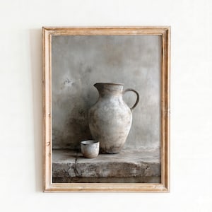 Puede incluir: Una lámina de arte enmarcada que muestra una jarra de cerámica grande y desgastada de color gris claro junto a una taza pequeña a juego. Los objetos tienen un acabado rústico y texturizado sobre una repisa de piedra y una pared neutra.