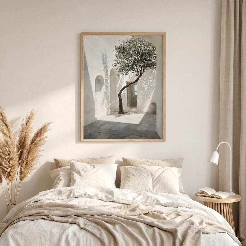 May include: A framed art print depicts a sunlit courtyard with a slender tree. The artwork features a white-walled building with arched doorways and a stone-paved floor. The tree's branches reach towards the light, creating shadows. The frame is a light wood color.