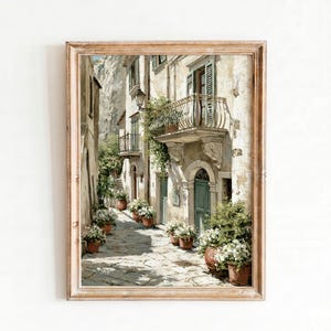 May include: A framed print of a European street scene. The artwork depicts a narrow cobblestone street lined with aged buildings, balconies, and potted flowers. The color palette is muted, with warm tones and a vintage aesthetic.