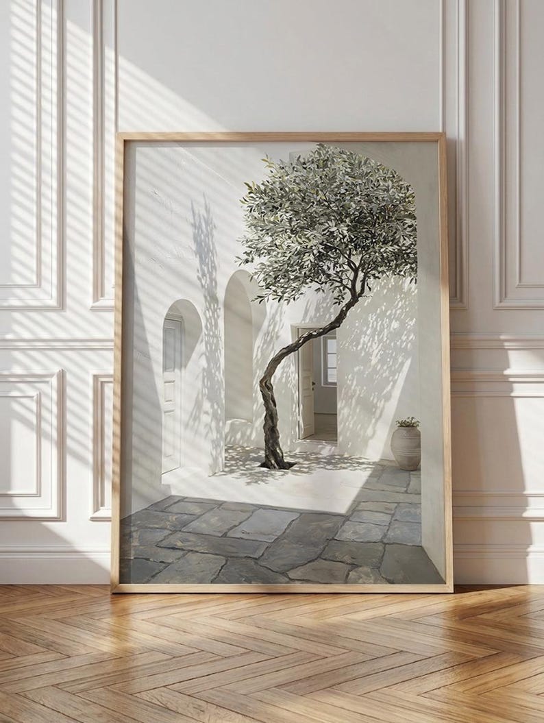 May include: A framed print depicts a sunlit courtyard with a slender olive tree. The tree's branches cast shadows on the white walls and stone floor. A wooden frame surrounds the artwork, which is displayed on a wooden floor.