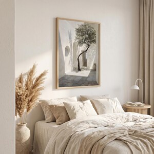 May include: A framed art print depicts a sunlit courtyard with a tree. The artwork features a white building with arched doorways and a stone-paved ground. The tree's branches reach towards the light, creating shadows. The frame is light-colored.