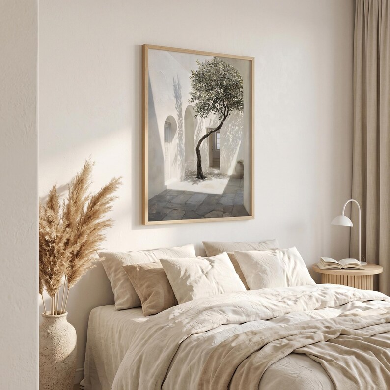 May include: A framed art print depicts a sunlit courtyard with a tree. The artwork features a white building with arched doorways and a stone-paved ground. The tree's branches reach towards the light, creating shadows. The frame is light-colored.