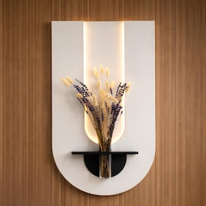 May include: A white wall-mounted decorative piece with a curved bottom and a vertical light strip. A black shelf holds a clear glass vase filled with dried lavender and wheat. The wall behind is wood-paneled.