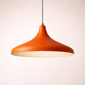 May include: Orange pendant light fixture with a wide, flared shade. The light hangs from a black cord and has a white interior. The fixture is a bright, solid orange color and is illuminated by a single bulb.