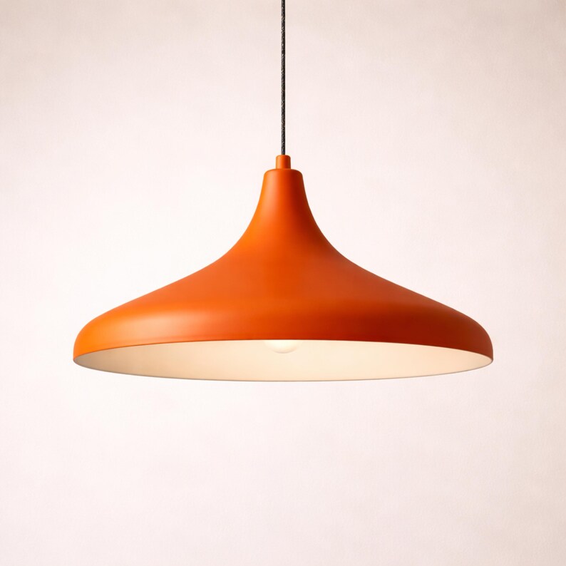 May include: Orange pendant light fixture with a wide, flared shade. The light hangs from a black cord and has a white interior. The fixture is a bright, solid orange color and is illuminated by a single bulb.