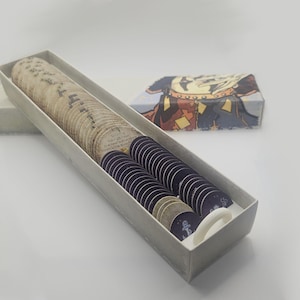 May include: A rectangular box filled with beige and dark blue circular tokens, possibly for a game. A smaller, patterned box sits nearby. The tokens are likely collectibles or game pieces.