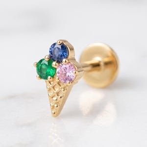 May include: A gold flat back stud earring shaped like an ice cream cone. The cone features a textured waffle pattern, while the top is adorned with three round faceted gemstones: a blue stone, a green stone, and a pink stone set in gold.