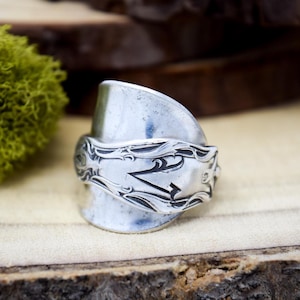 May include: A silver-toned ring crafted from a repurposed spoon, featuring a wide, curved band that wraps around the finger. The surface of the ring displays intricate, embossed scrollwork patterns along the handle section, with a polished finish.