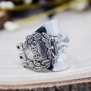 May include: A silver-toned metal ring crafted from a repurposed spoon handle. The ring features a prominent circular emblem embossed with the word YEARS, surrounded by a detailed oak leaf wreath pattern and decorative etched line accents.
